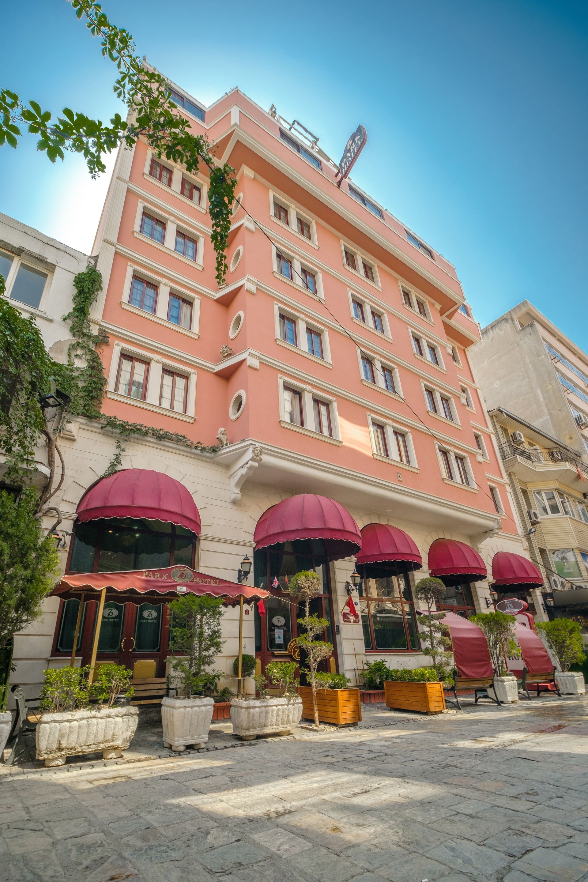 Oğlakçıoğlu İzmir Park Hotel building features red awnings, stone-paved walkways, and potted plants in an outdoor setting.