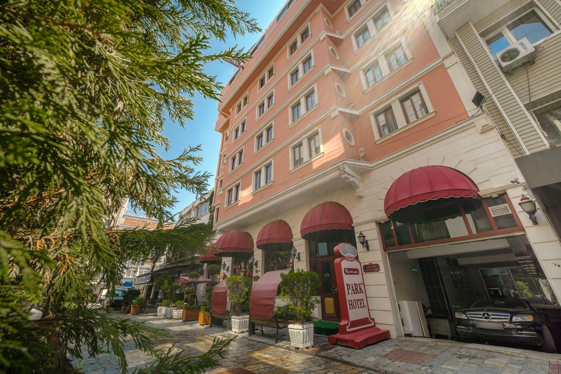Oğlakçıoğlu İzmir Park Hotel building with red awnings over the ground-level entrances, viewed from a leafy street.