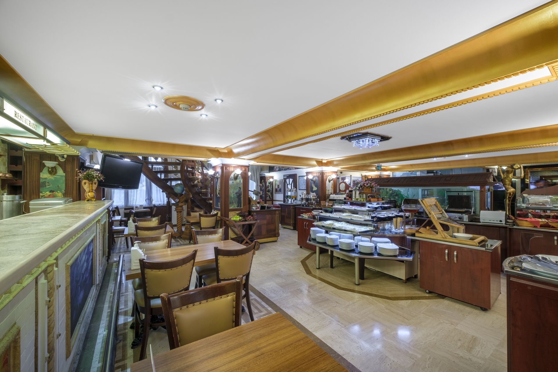 The interior of an elegant, well-lit breakfast buffet area with tables, chairs, and a serving counter with stacked plates.