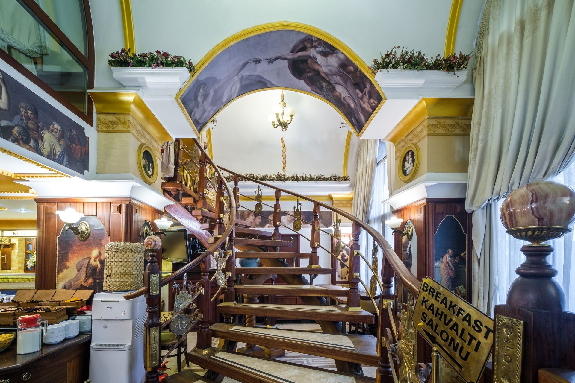 A wooden spiral staircase leading to a mezzanine, framed by an archway with a mural of the Creation of Adam in a hotel.