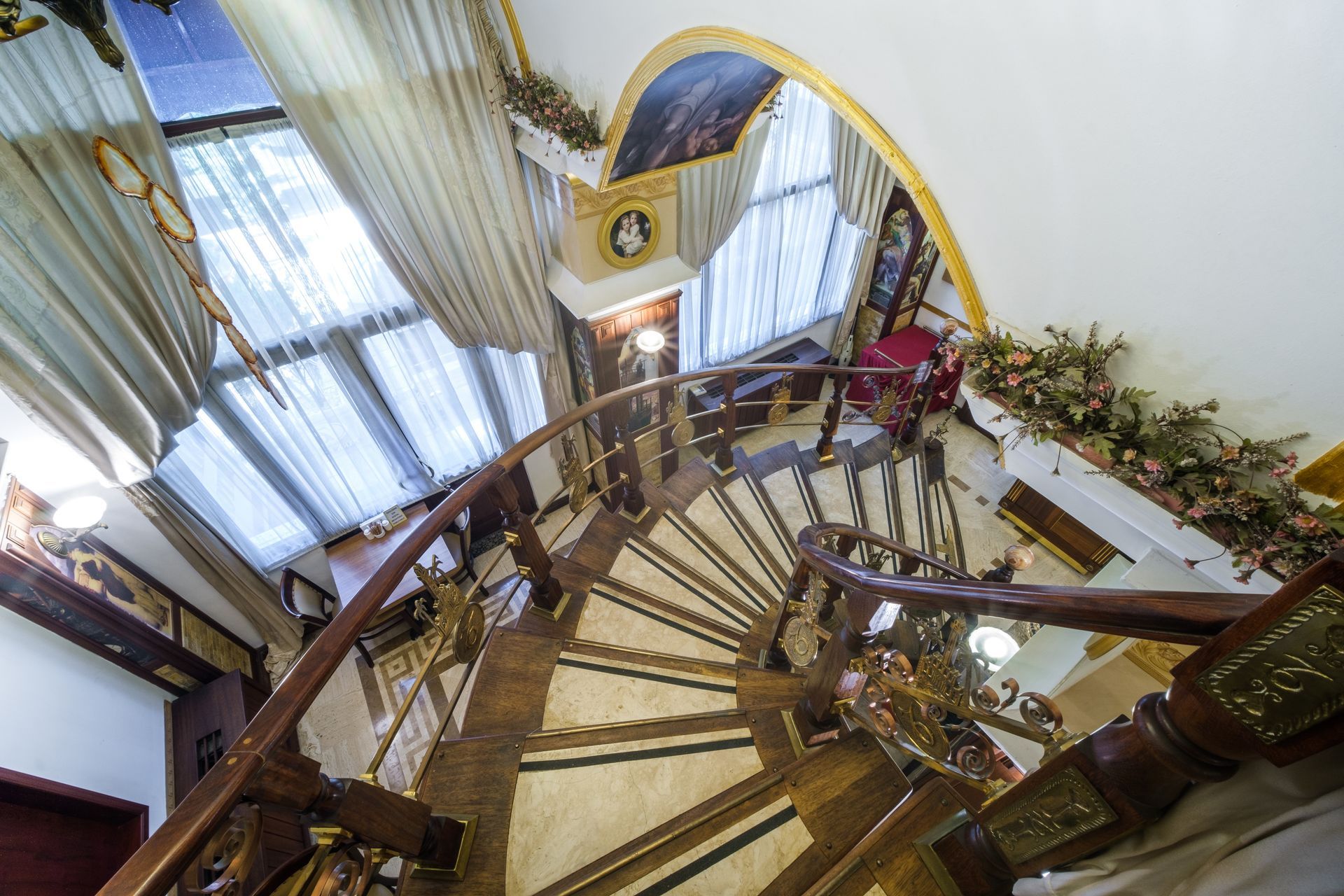 High-angle view of a curved wooden staircase with ornate railings, leading down into a bright room with tall windows.