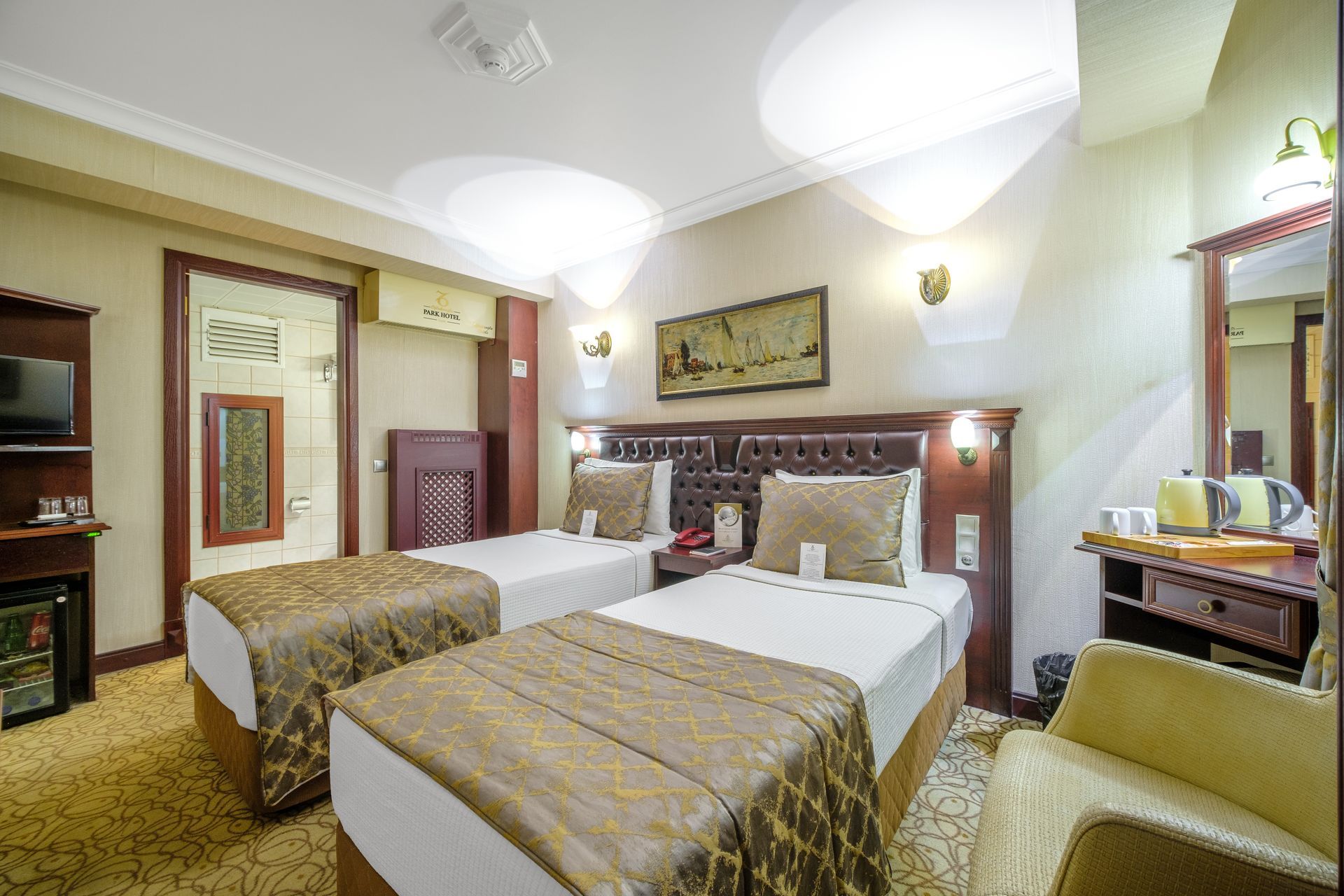 İzmir Park Hotel's room with two twin beds featuring tan patterned runners, a desk, a mirror, and a small refrigerator.