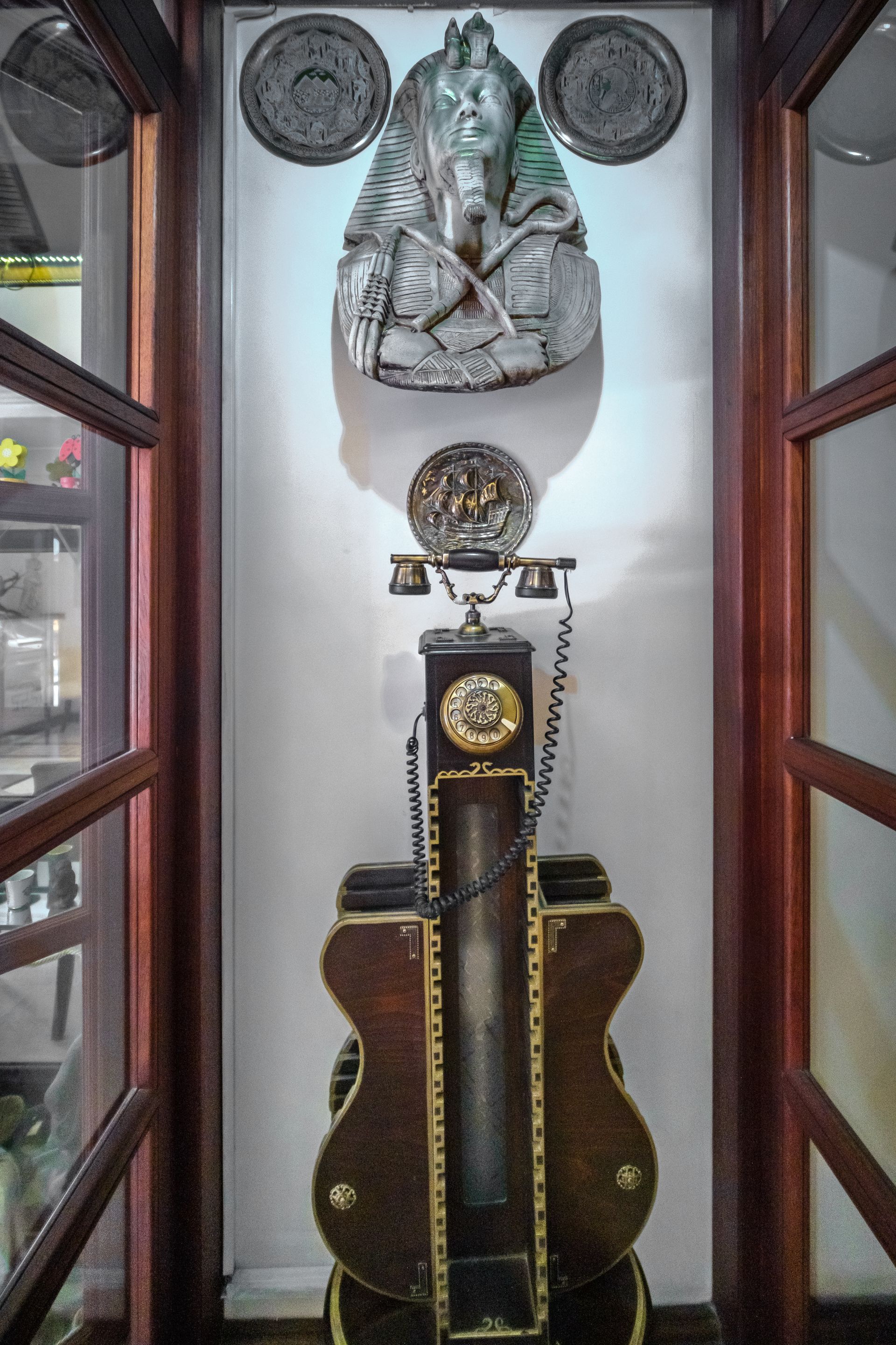 A decorative wall piece featuring a Pharaoh bust above an ornate, violin-shaped vintage telephone in a glass display.