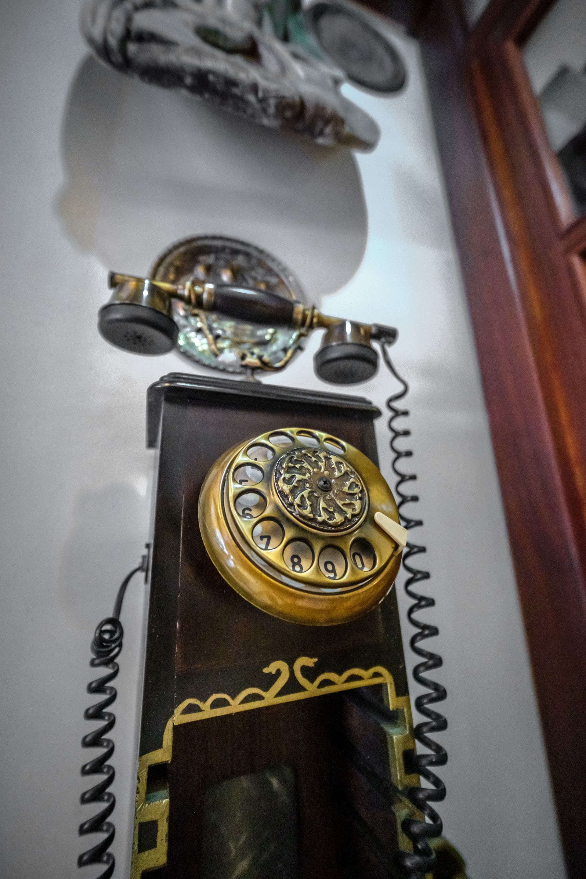 A vintage, wall-mounted rotary telephone with a wooden body, gold-colored dial, and curly black cord on a white wall.