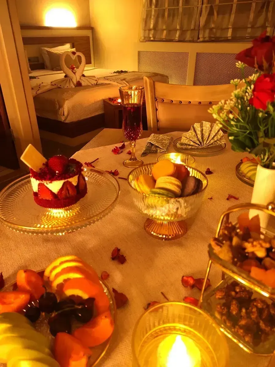 A romantic setup with a strawberry dessert, macarons, and fruit on a candlelit table, with a bed visible in the background.
