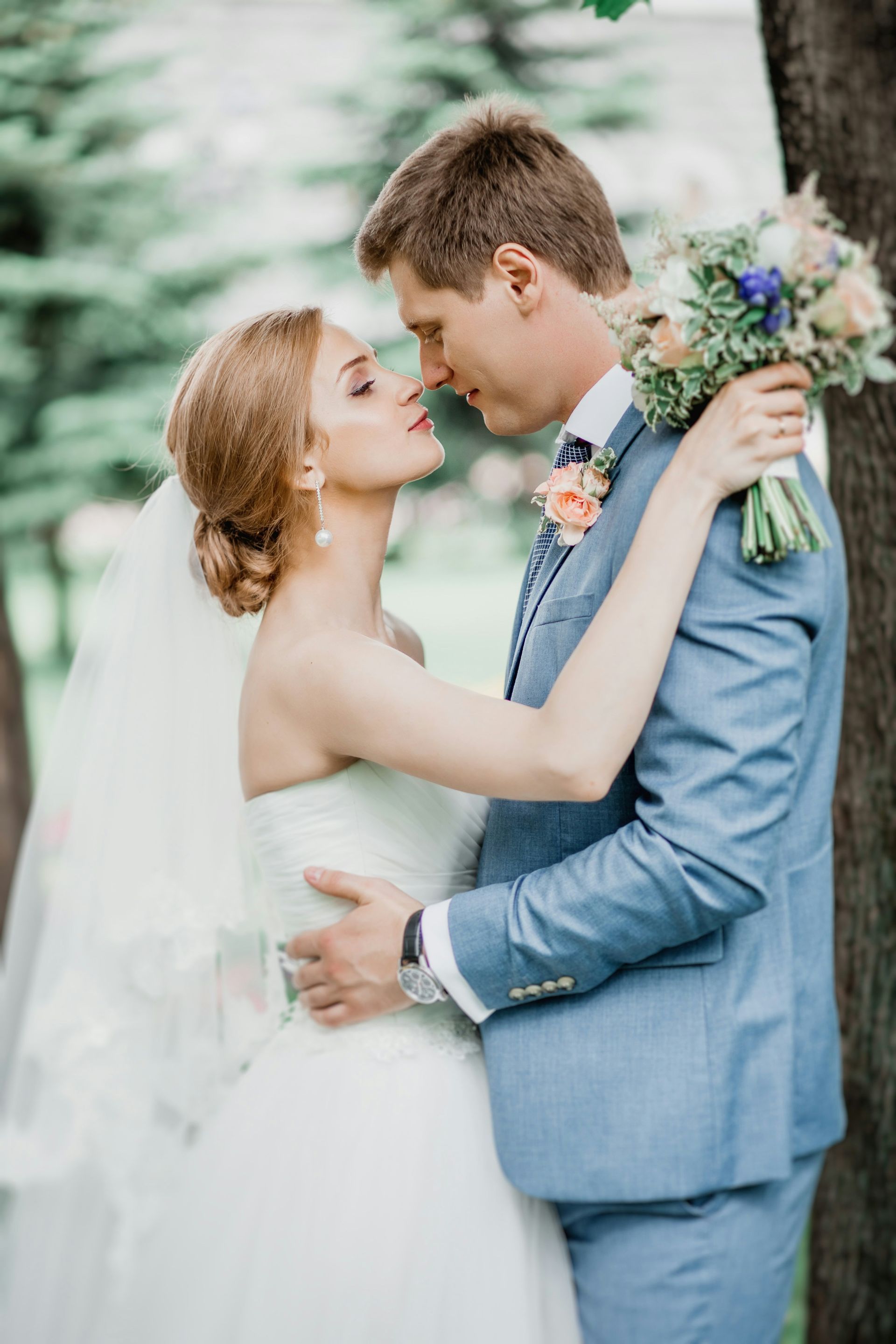 A bride and groom are kissing in a park while the bride is holding a bouquet of flowers.