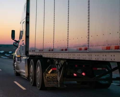 A semi-truck travels on a highway at dusk, with the sky showing orange and purple sunset hues.