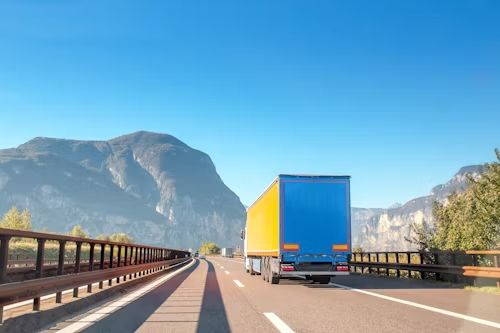 Semi-truck on a highway with blue sky, mountains in the background. The trailer is blue and yellow.