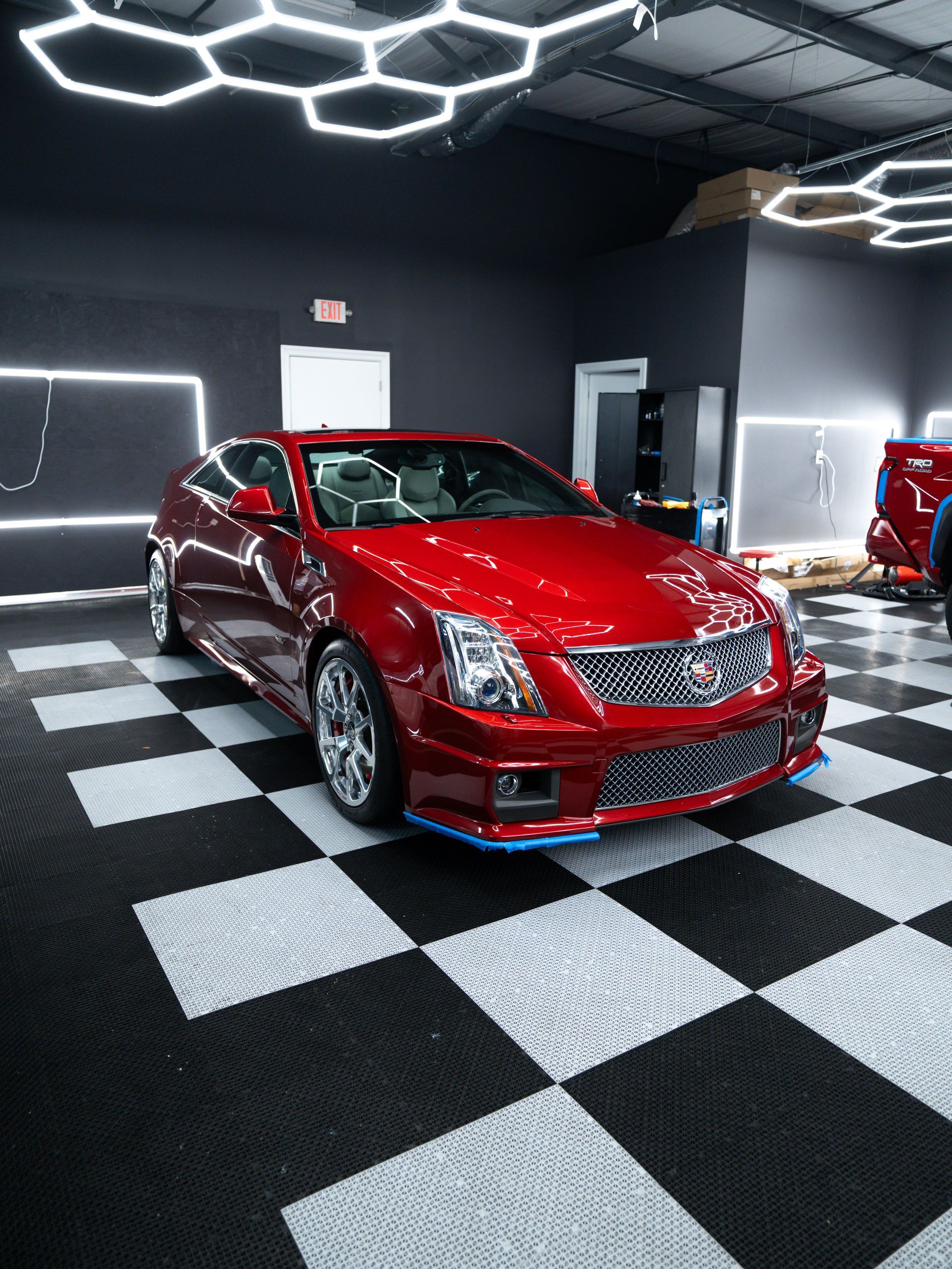 Red sports sedan in a glossy showroom with black-and-white checkered floor and hexagonal ceiling lights