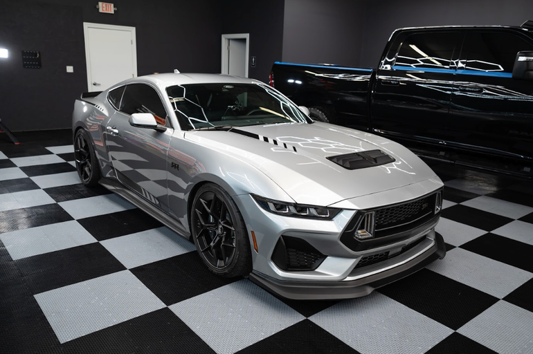Silver sports car in a checkered-floor showroom, parked beside a black vehicle and a large truck.