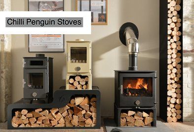 Chilli Penguin Stoves: Display of three wood-burning stoves with stacked firewood; one stove has a lit fire.