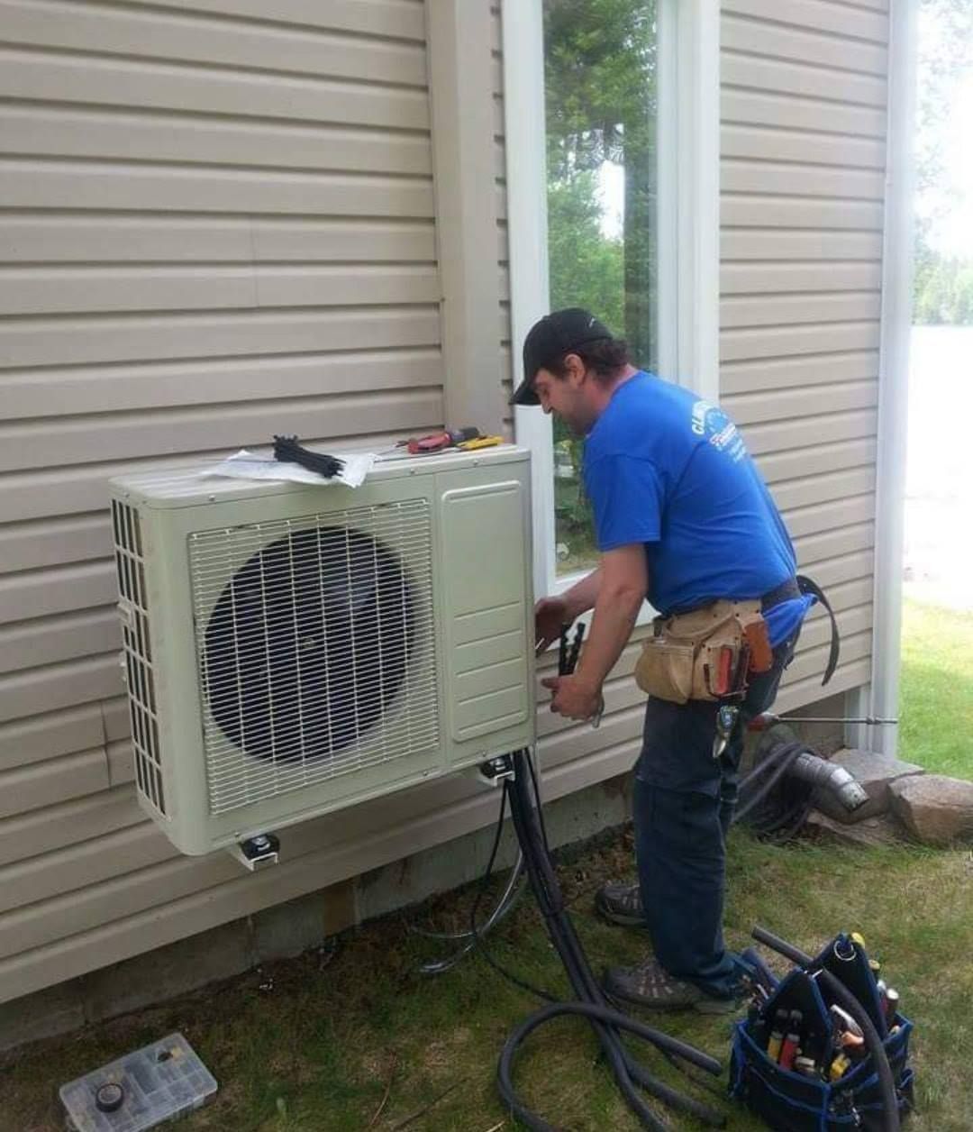 Un homme en chemise bleue travaille sur un climatiseur sur le côté d'une maison.