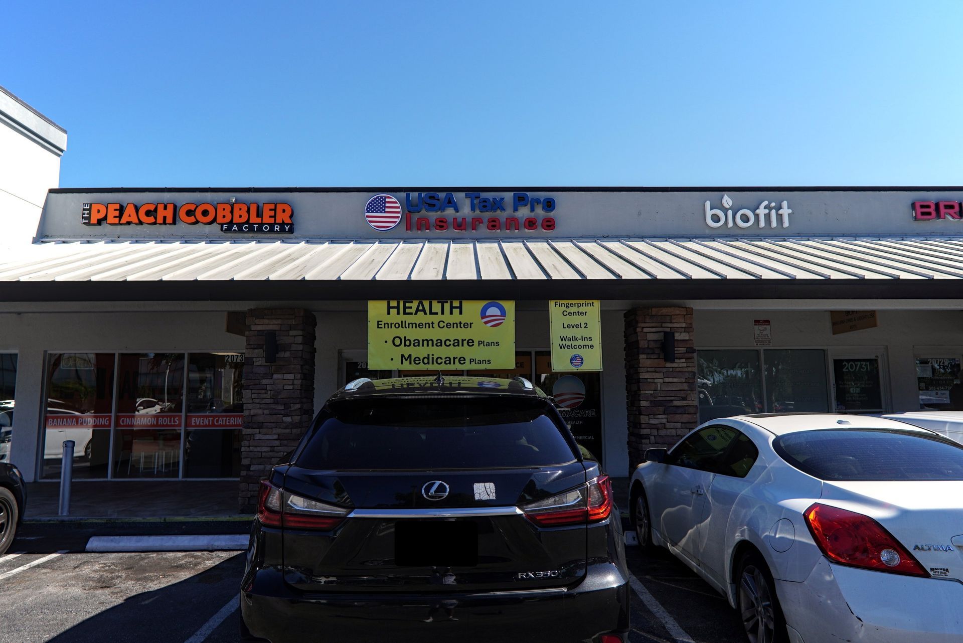 Exterior of a strip mall with businesses including a tax service, gym, and restaurant. Cars are parked out front.