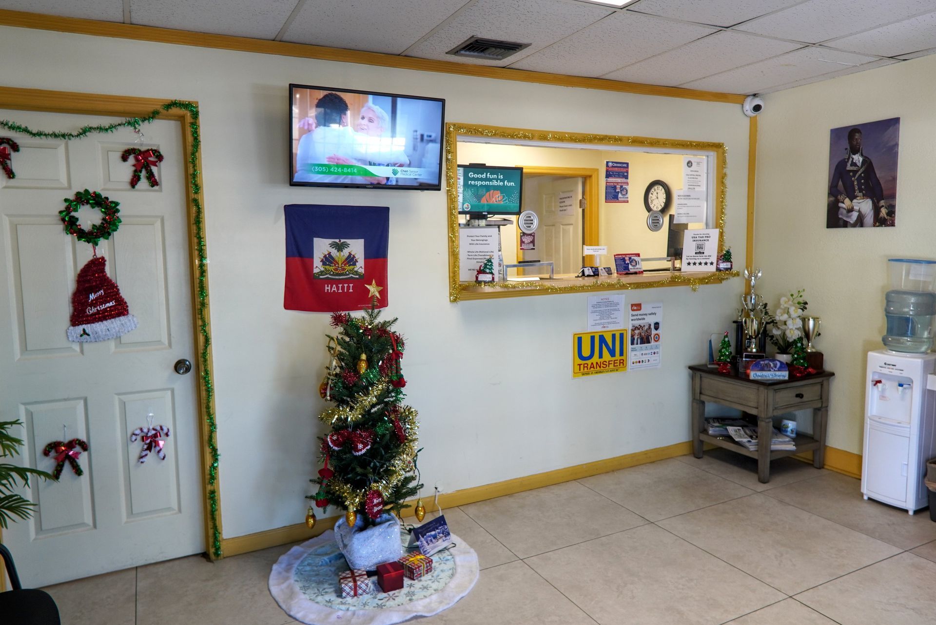 Interior of a waiting room; Christmas decorations; Haitian flag; receptionist window; TV on wall; water dispenser.