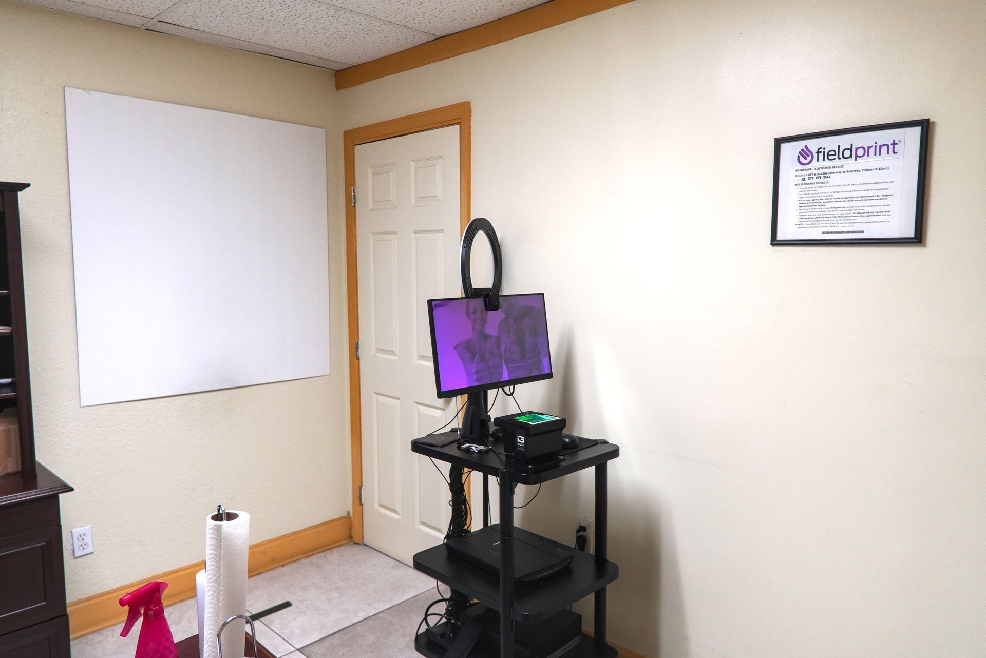 A room with a whiteboard, a desk holding a computer and a ring light, and a door.