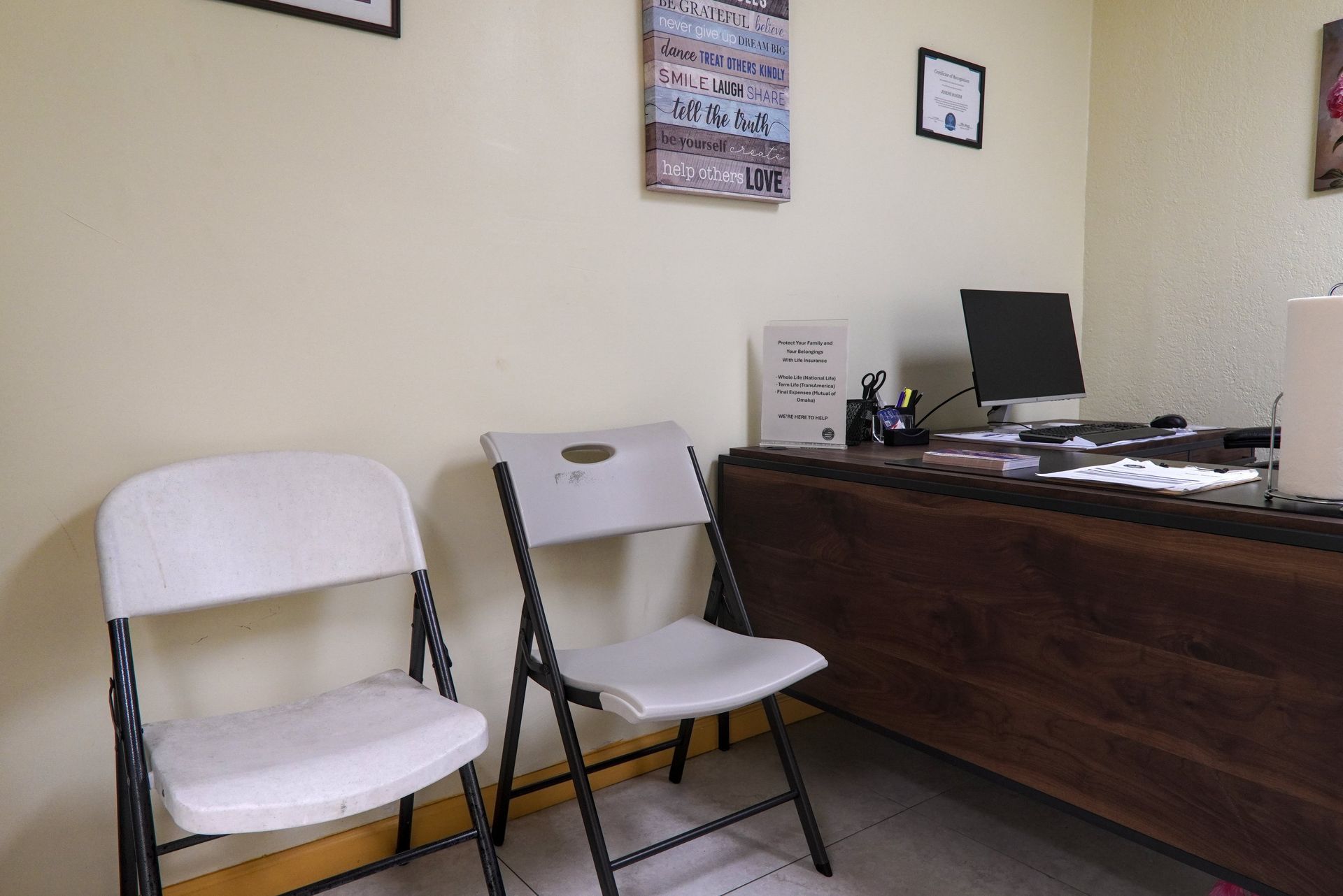 Two white folding chairs in a waiting area with a desk, computer, and wall decor.