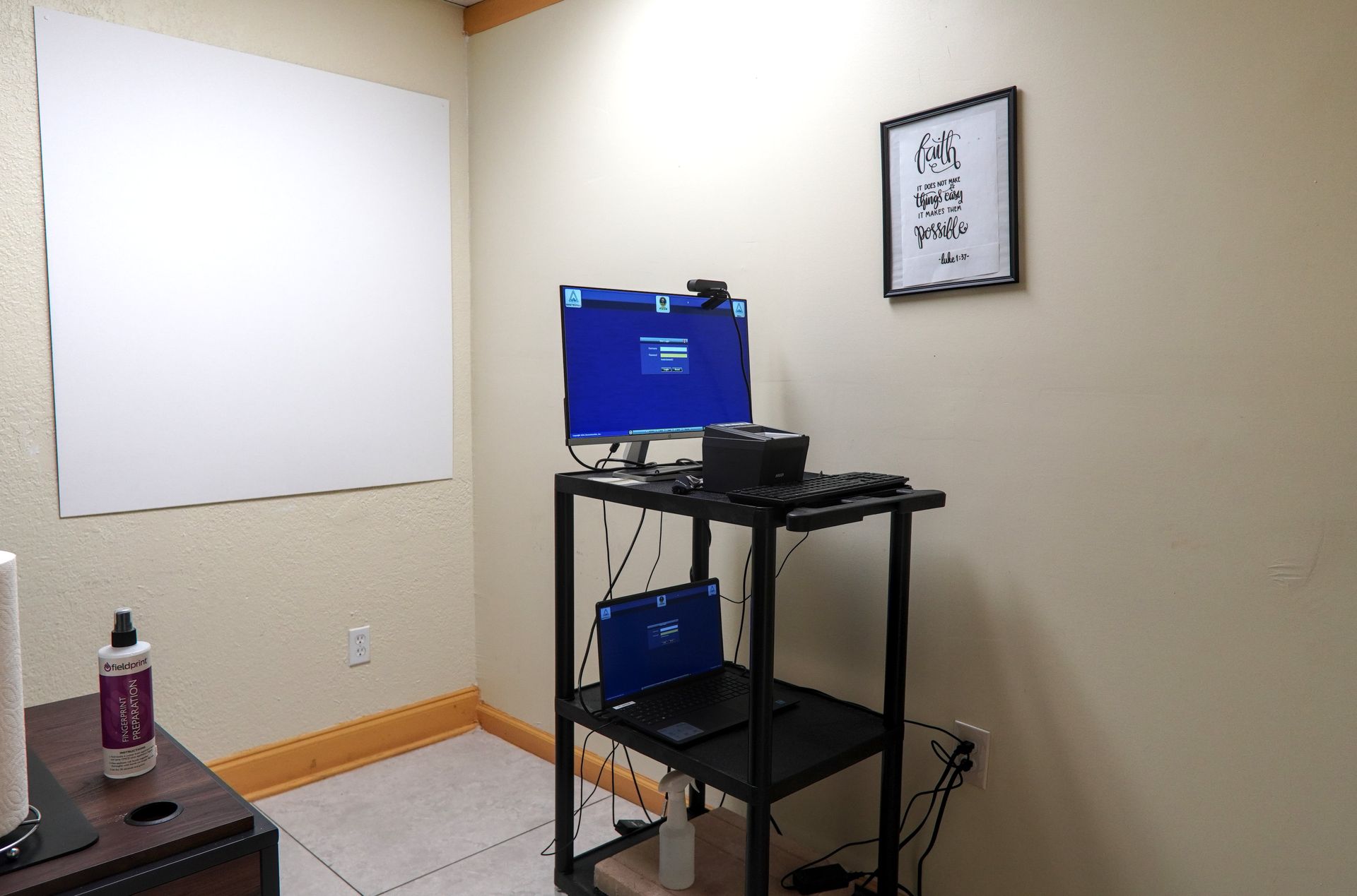 A small office with a whiteboard, desk with computer and laptop, and framed sign on the wall.