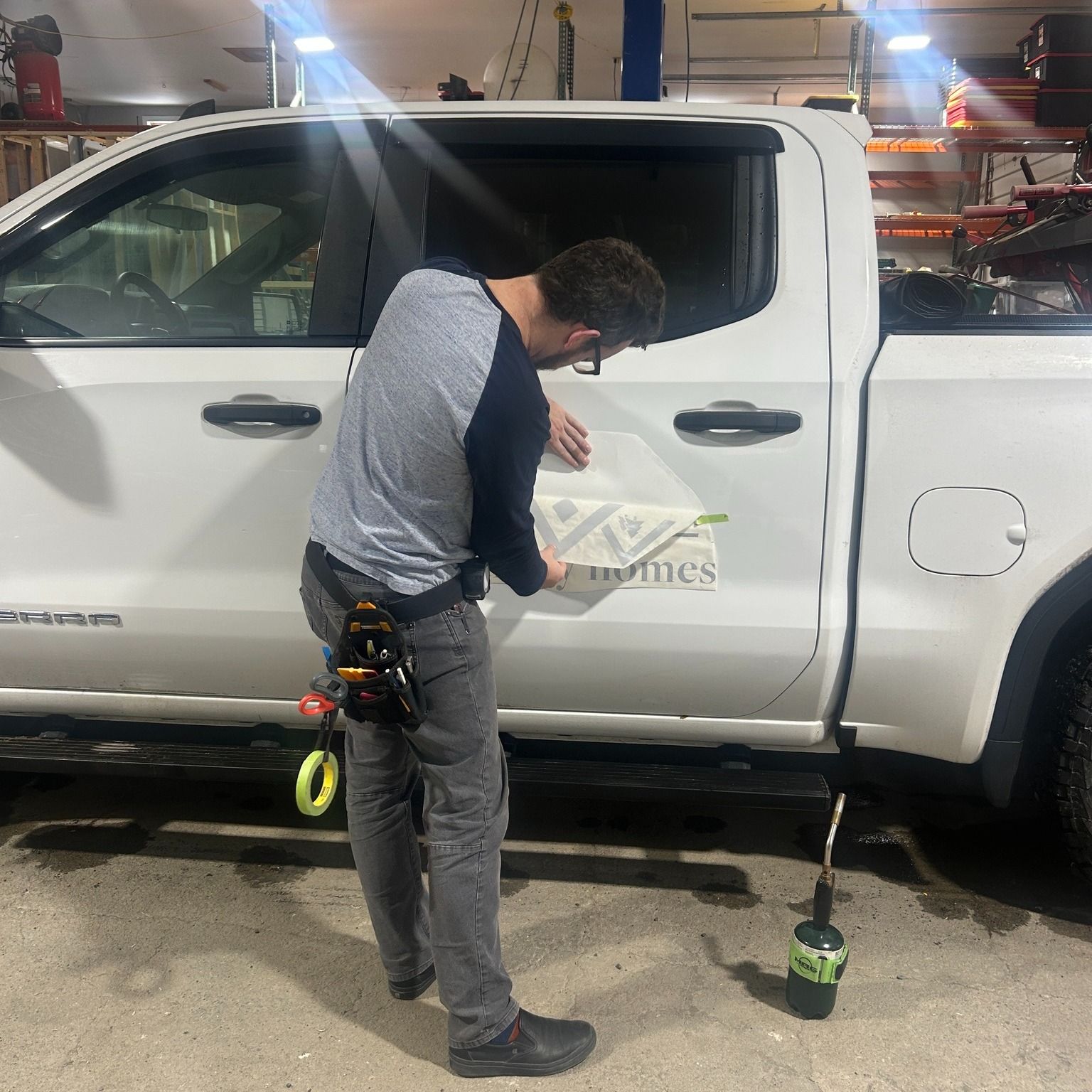 Man applying a logo sticker to the side of a white pickup truck. He's in a garage-like setting with tools nearby.