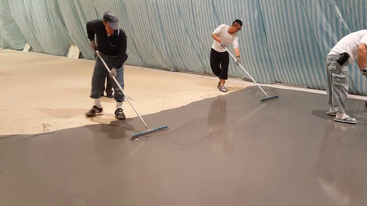 Three people applying a smooth, gray coating to a floor with long-handled squeegees in an indoor setting.
