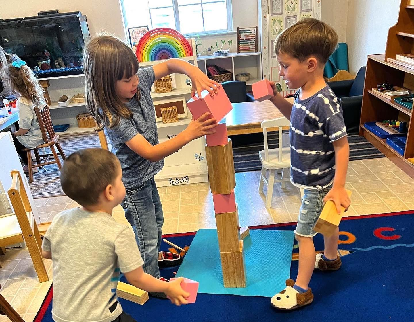 Three children build a tall tower using pink and wooden blocks on a blue rug in a classroom.
