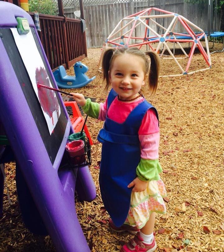 A smiling child in a blue smock paints on an outdoor easel in a playground with a dome climber in the background.