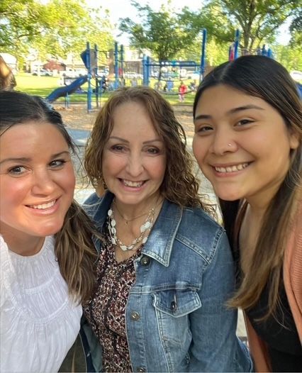 Three smiling people pose for a selfie outdoors at a playground with blue climbing equipment in the background.