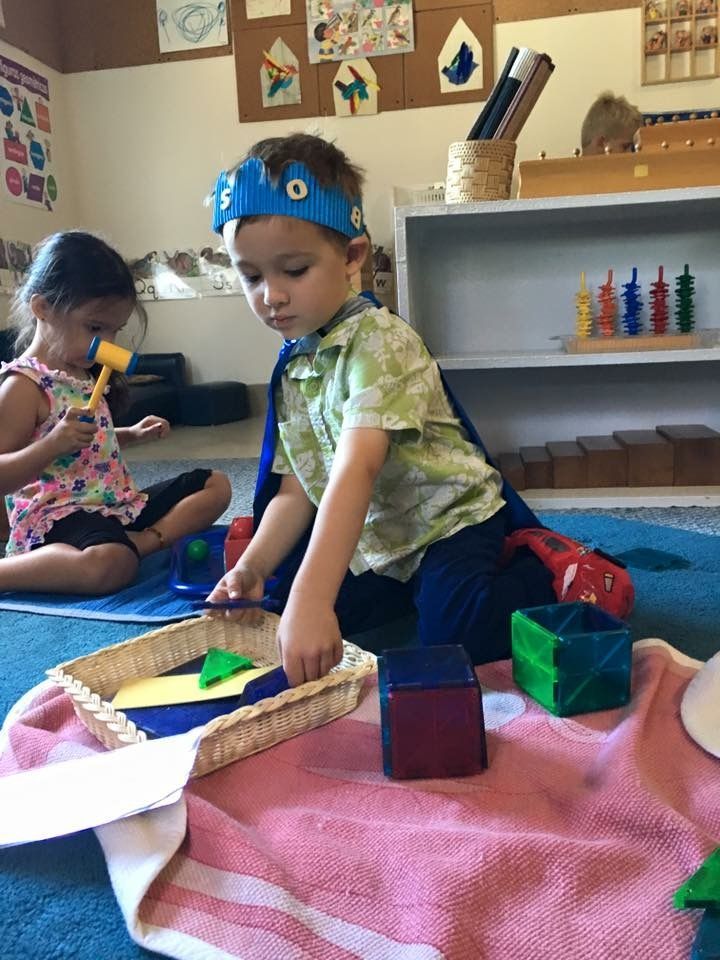 Two children sit on a rug playing with colorful blocks and shapes in a brightly lit classroom.