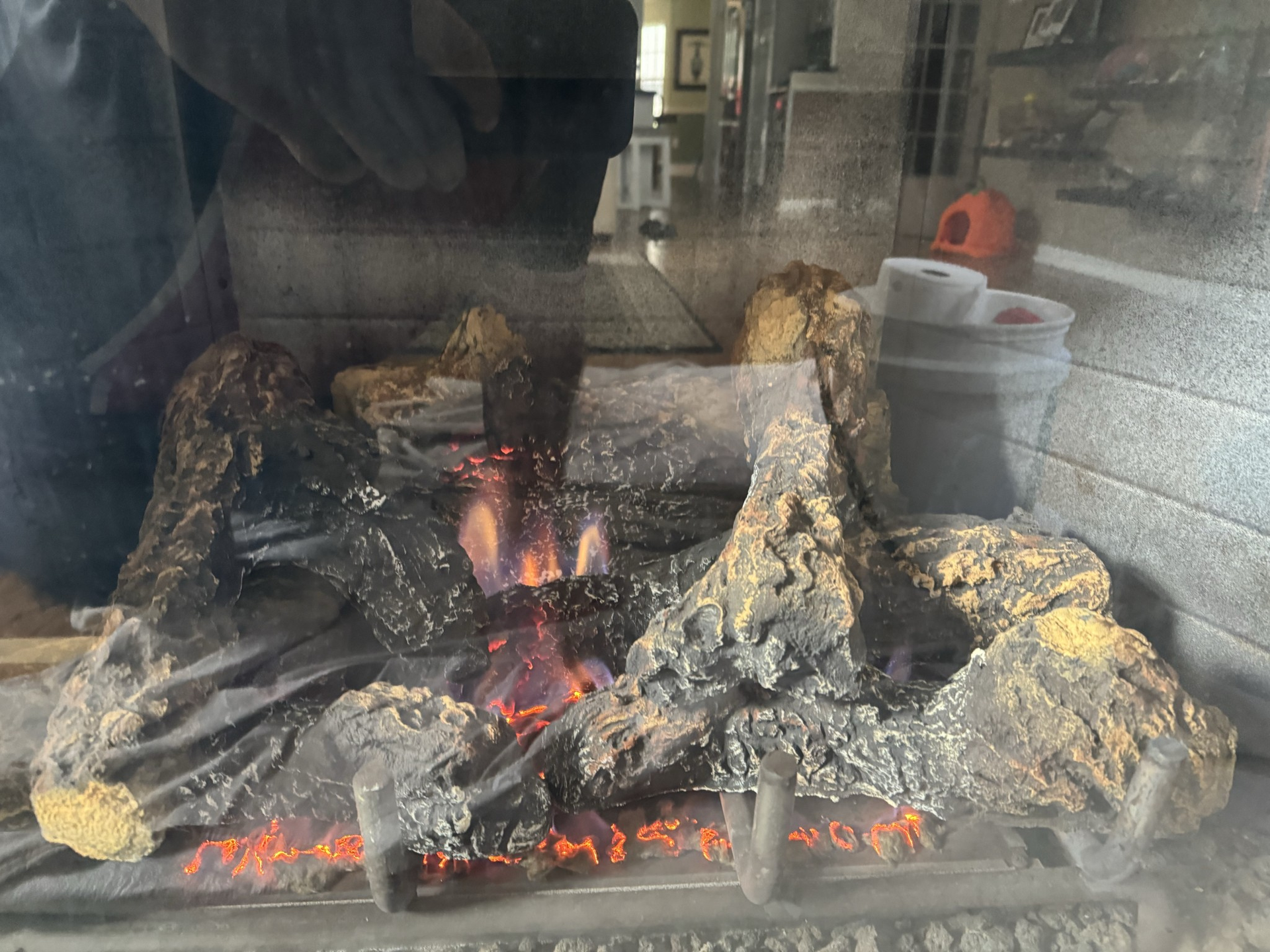 A lit gas fireplace with artificial logs, viewed through glass. Flames glow, reflections visible.