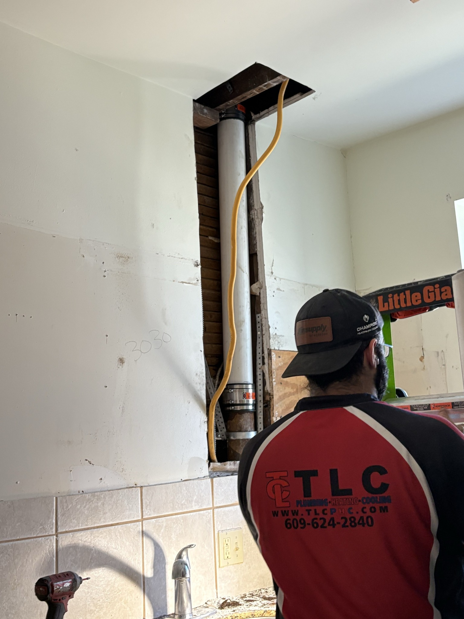 Man looking at exposed pipes inside a wall. Yellow electrical wire runs along white pipes.