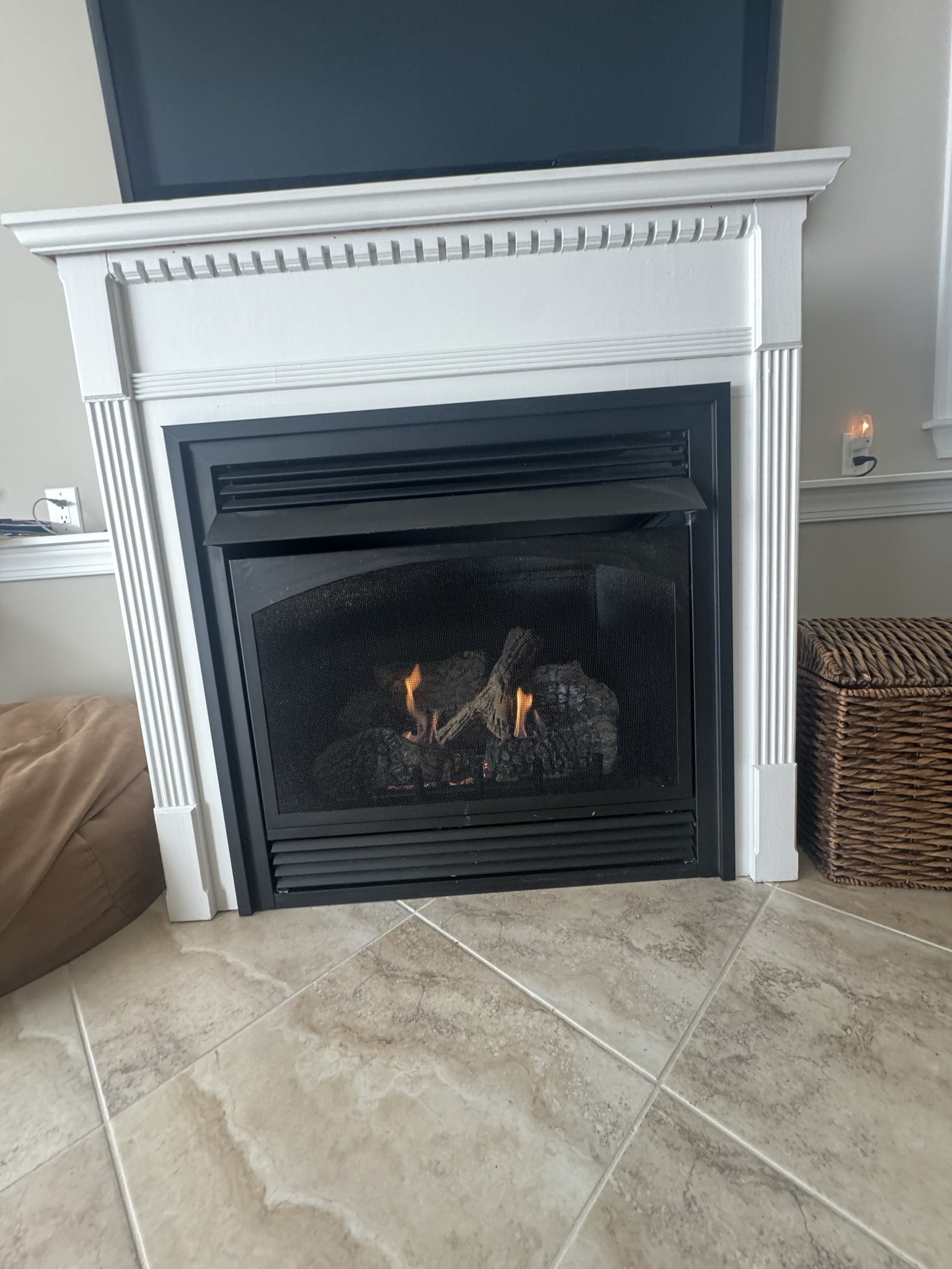White fireplace with black mesh screen, flames burning. Beige tile floor.