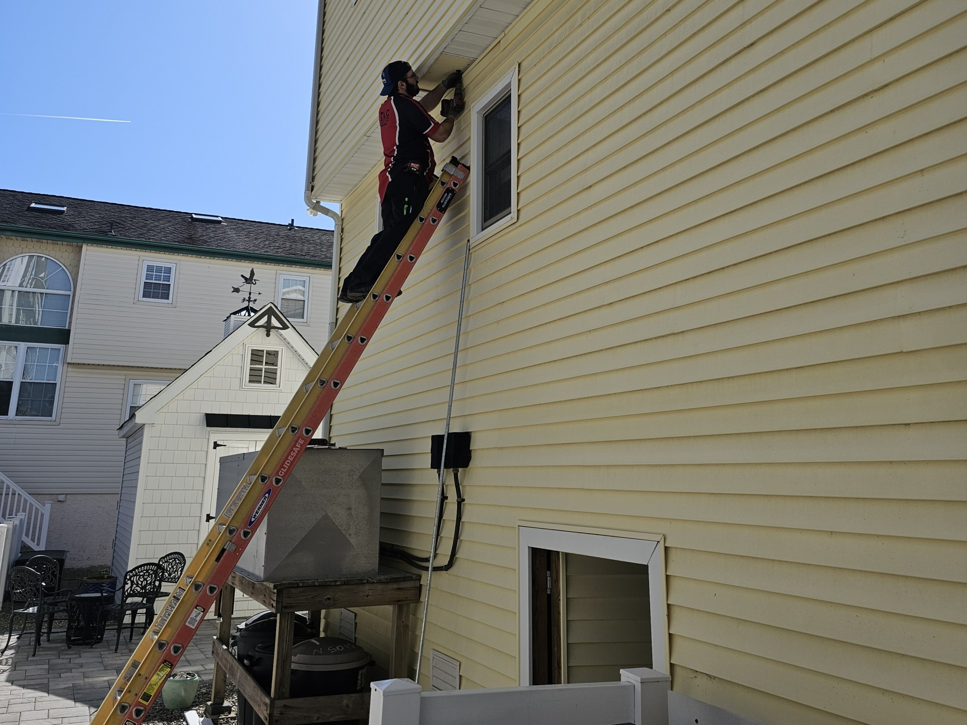 Person on a ladder, working on the side of a yellow house on a sunny day.