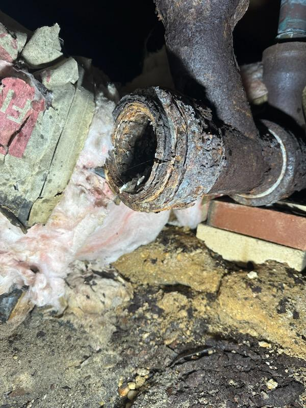 Corroded metal pipe connection with visible rust and decay. Surrounded by insulation, brick, and soil.