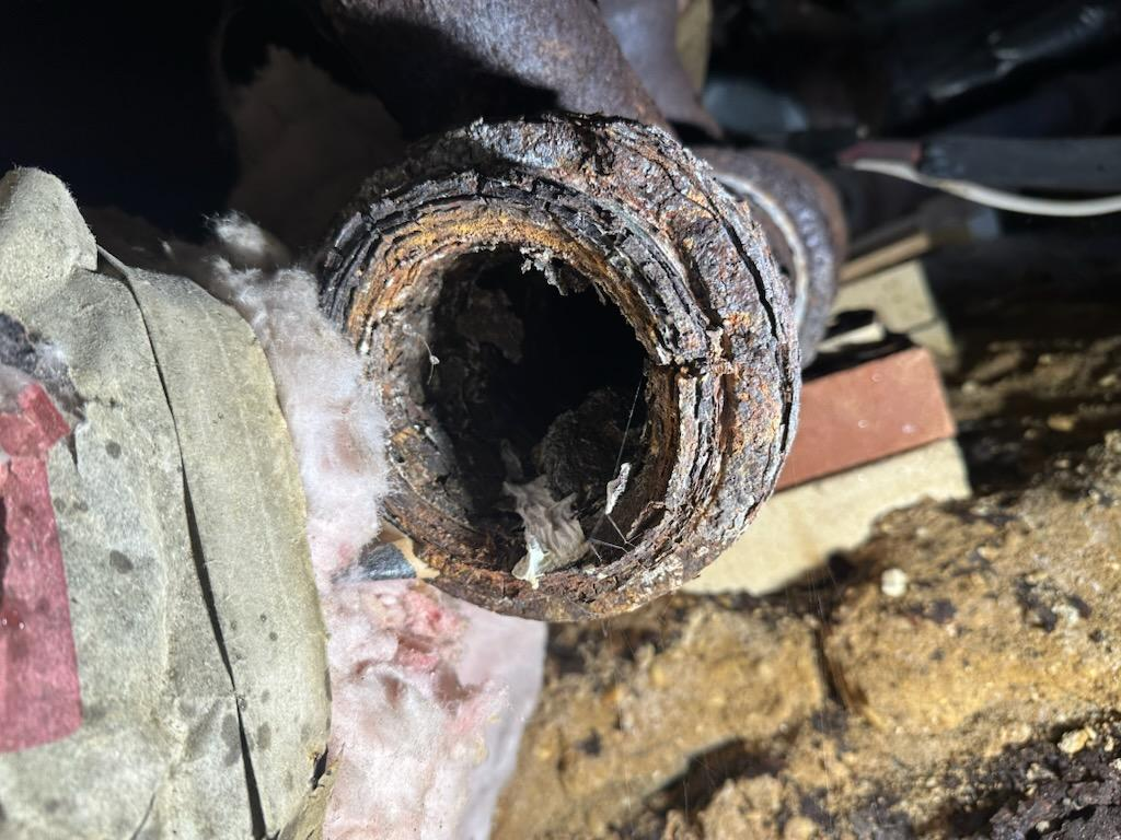 Severely corroded pipe with a large hole, revealing internal layers and debris; in a dim, cluttered space.