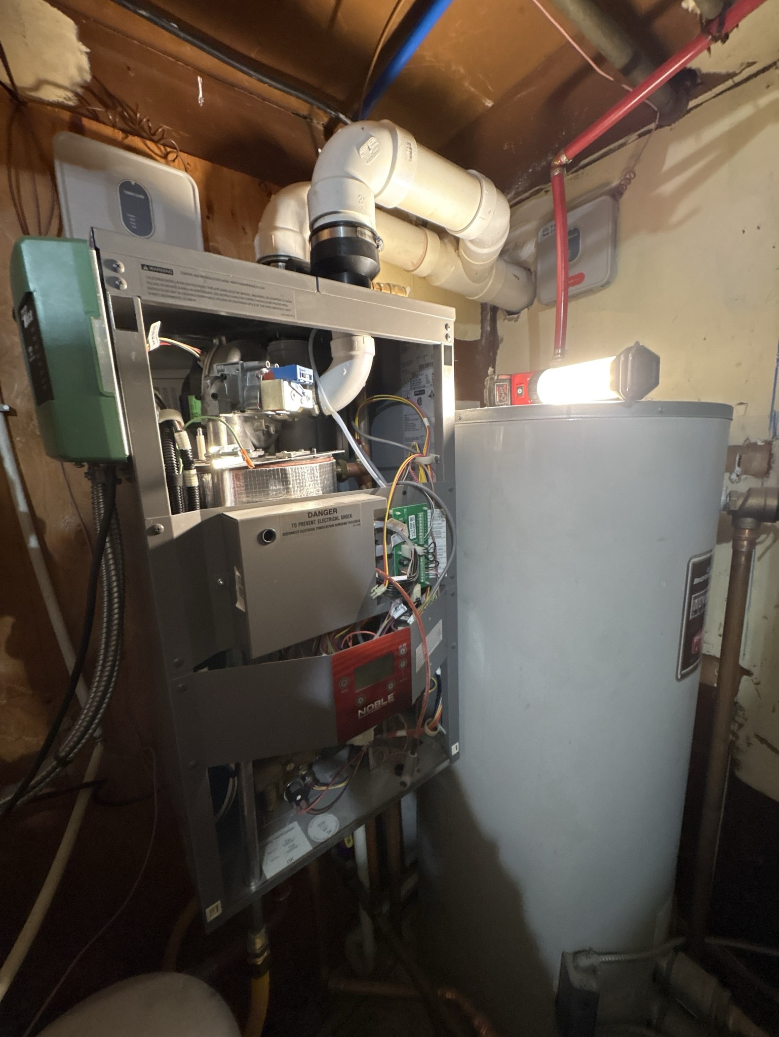 Furnace and water heater in a utility room. Grey furnace with exposed components, connected to a white pipe. A tall, silver water heater stands beside it.