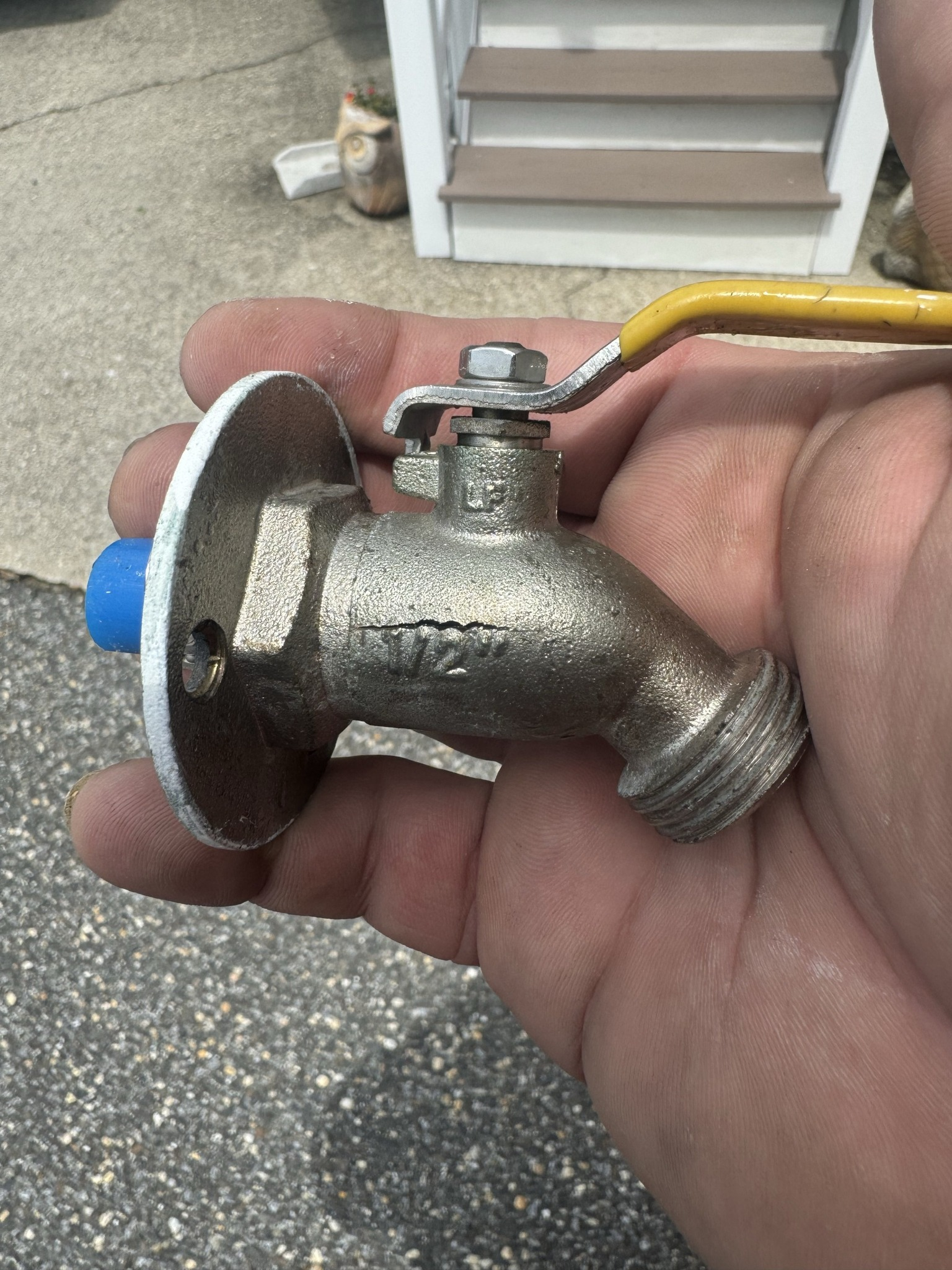 Hand holding a brass outdoor faucet with yellow handle; blue cap on opposite side; crack visible.