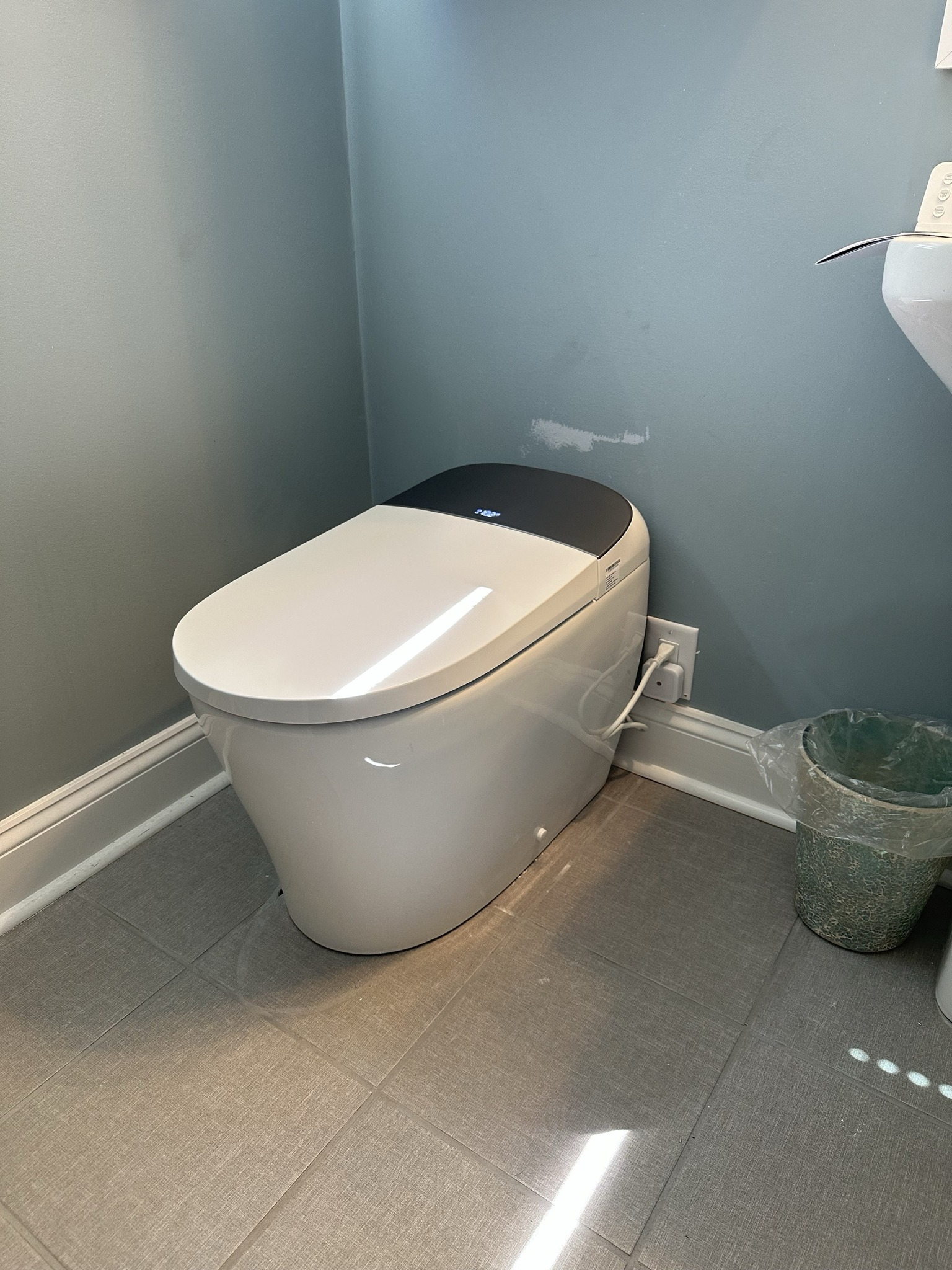 White modern toilet in a small bathroom with gray walls, flooring, and a green waste bin.