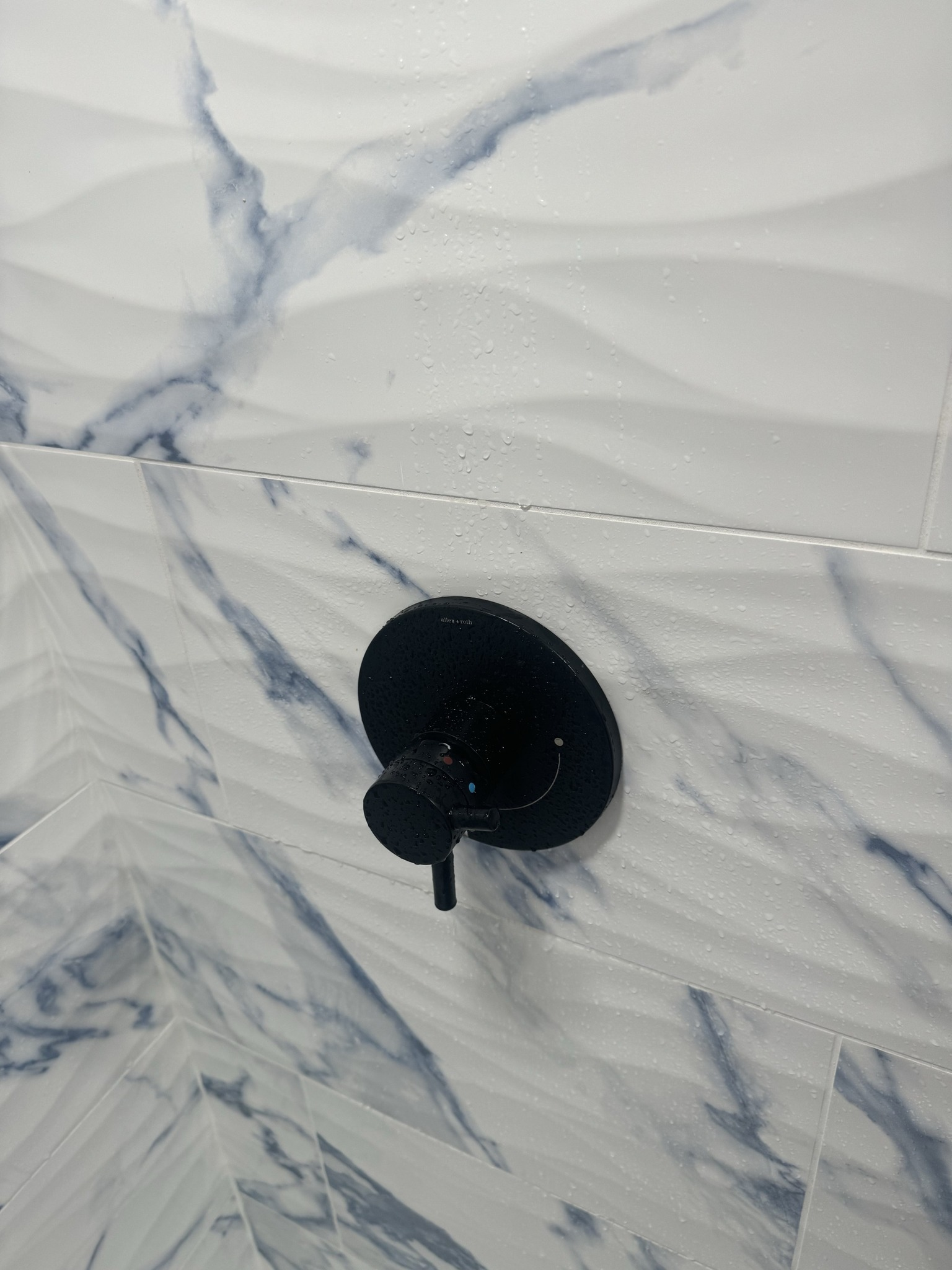 Black shower valve on marble-patterned tile wall with water droplets.
