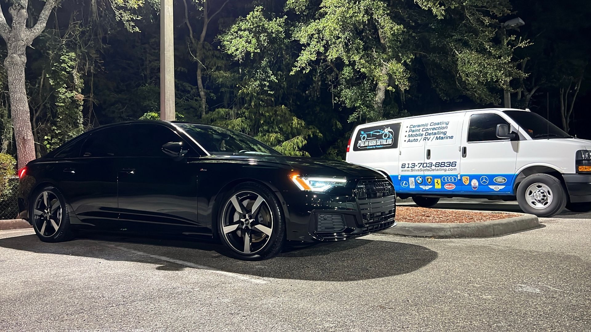 A black car is parked next to a white van in a parking lot at night.