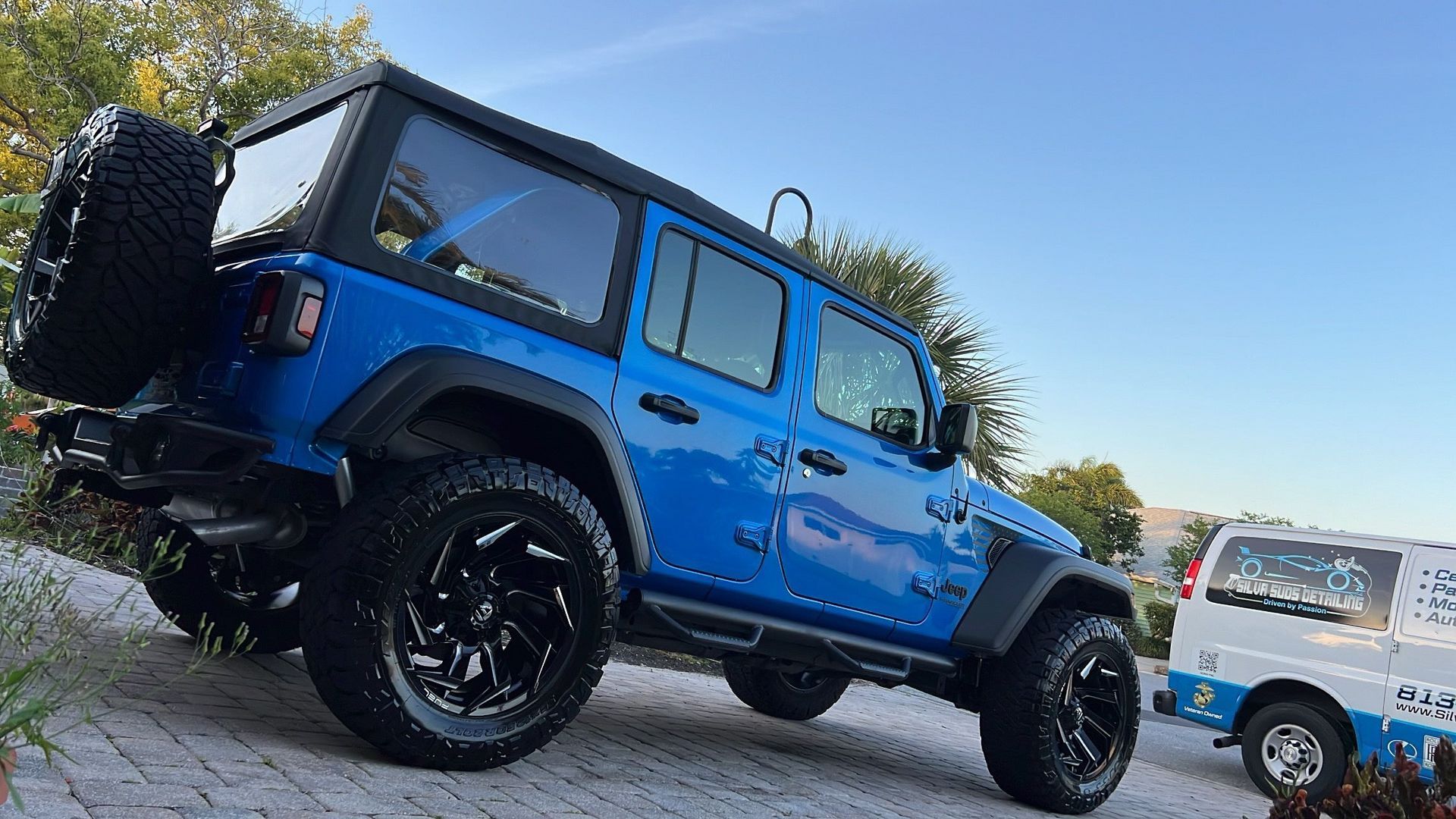 A blue jeep is parked on a brick driveway next to a white van.