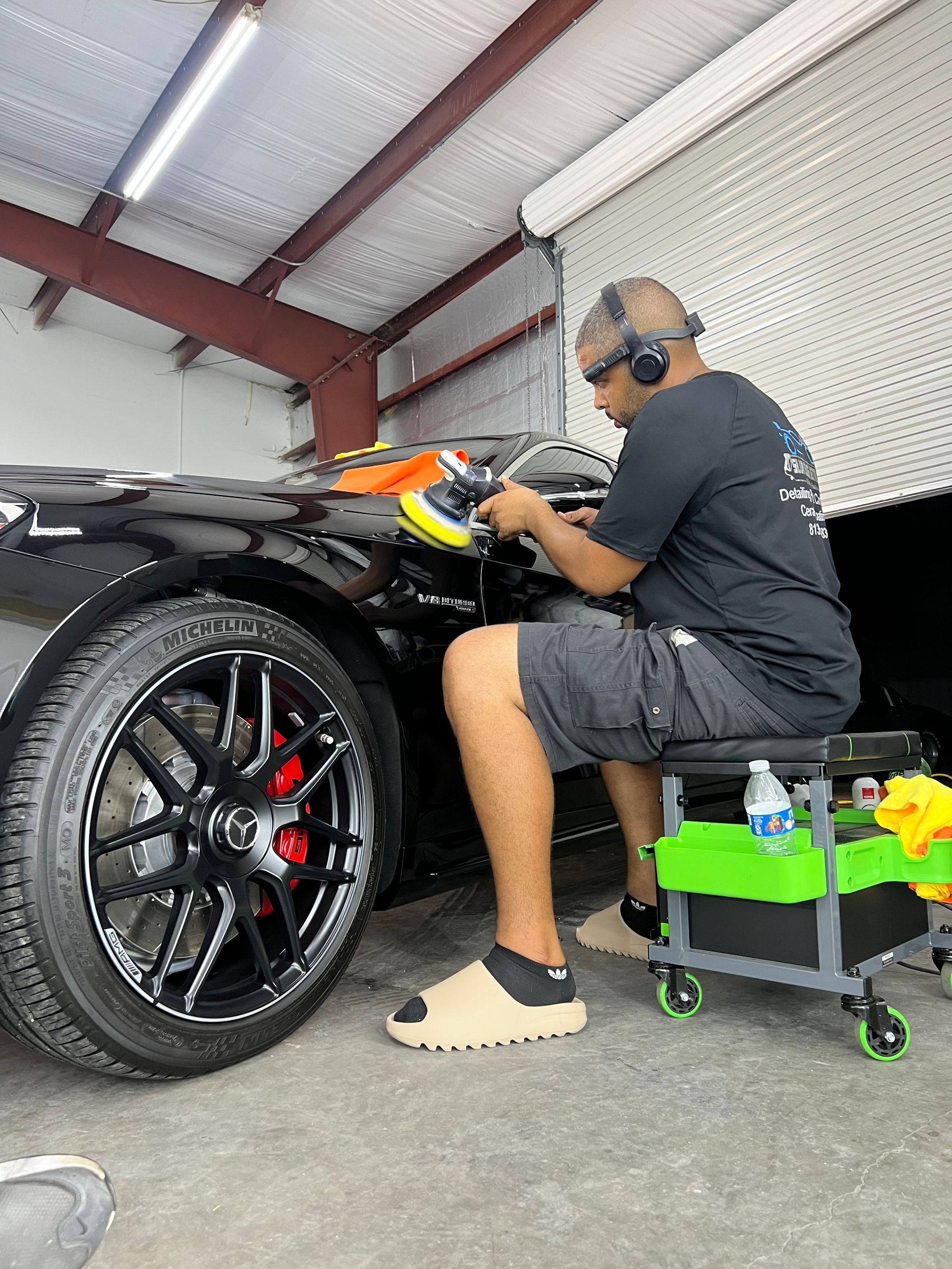 A man is sitting on a stool polishing a car in a garage.