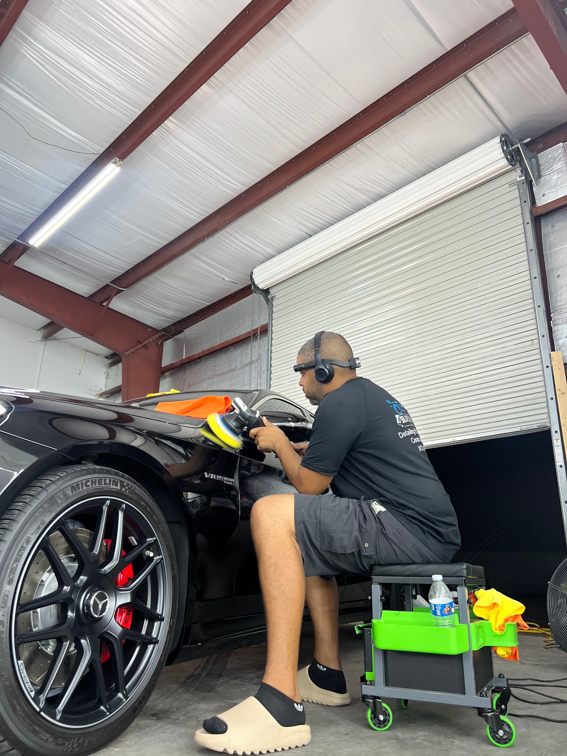 A man is sitting on a stool polishing a car in a garage.