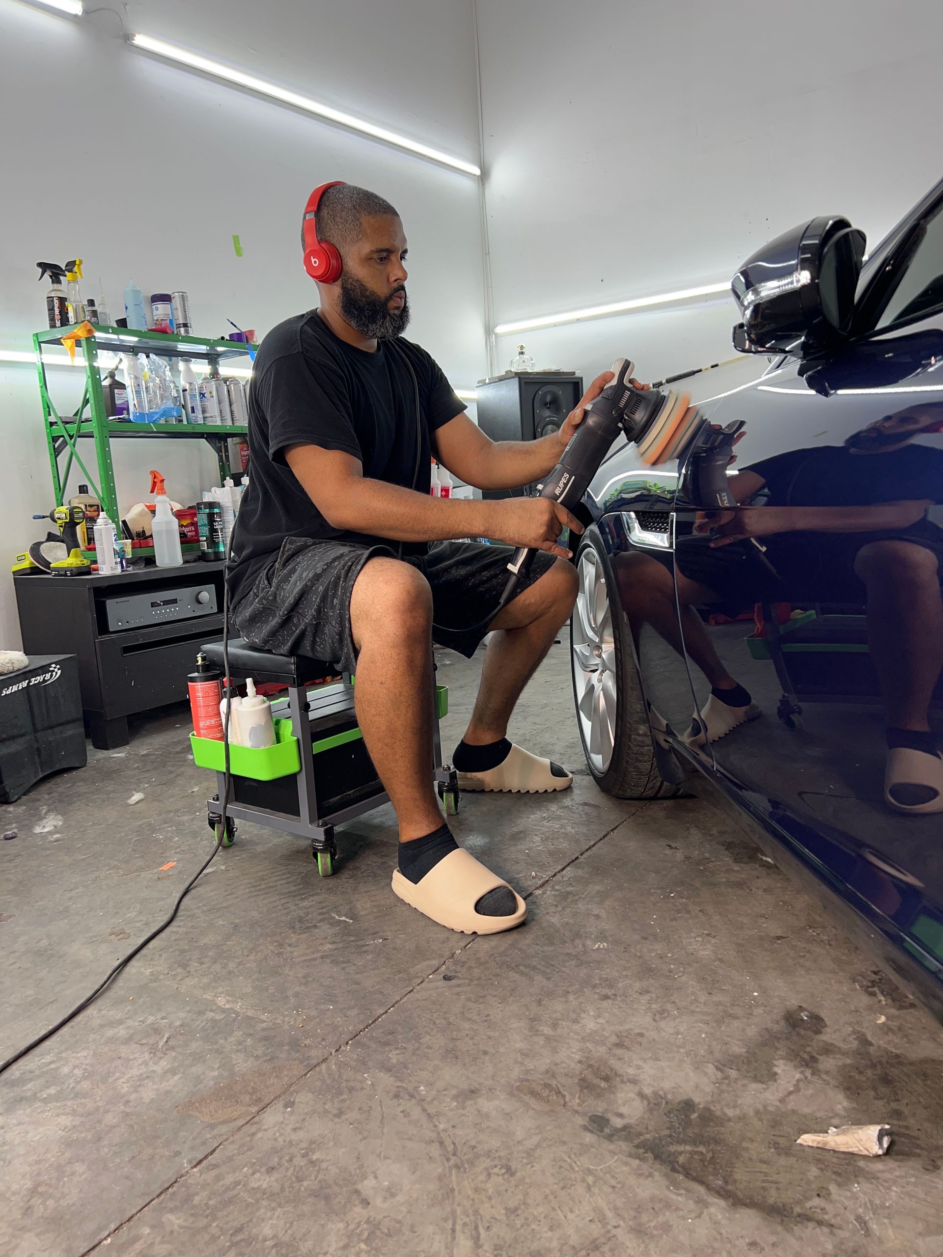 A man is sitting on a stool polishing a car in a garage.