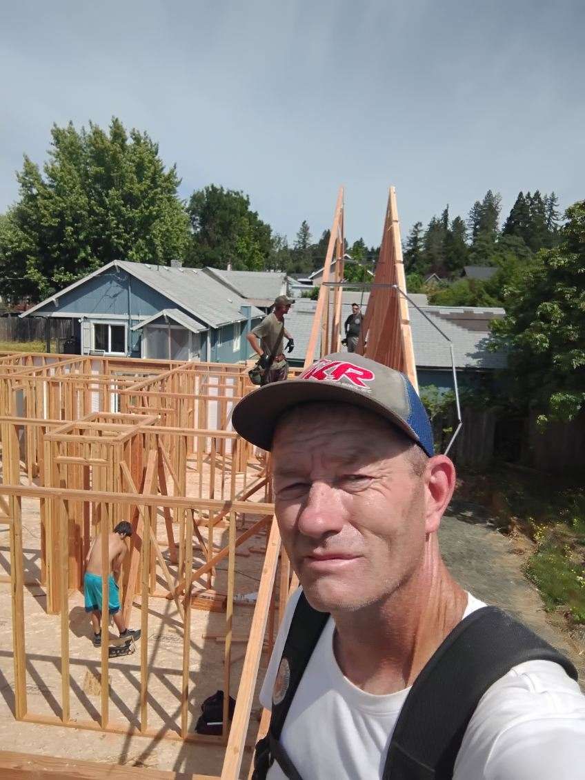 Man takes selfie on a construction site. Workers building wooden frames, blue house in background.