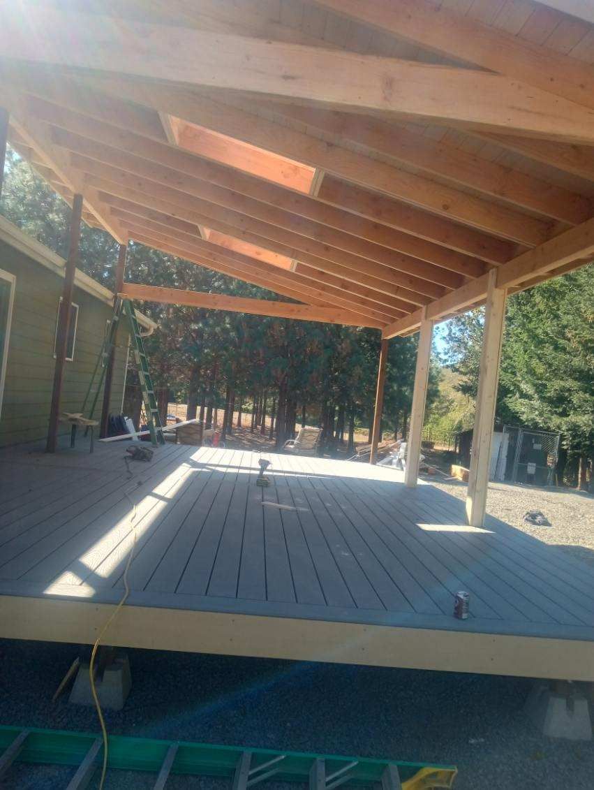 Wooden deck with a sloped roof, extending from a house into a wooded area.