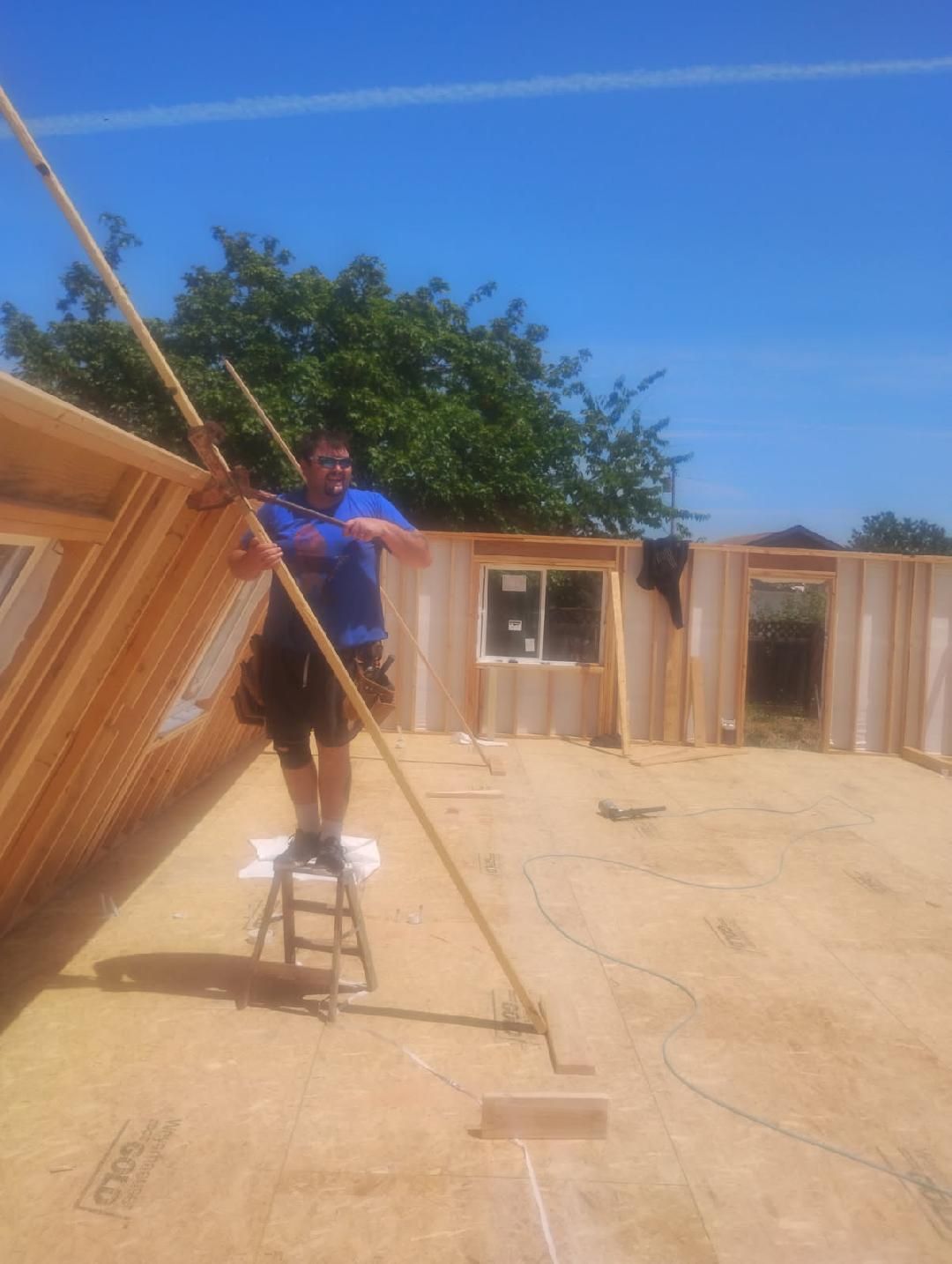 Construction worker on a stool using a pole to prop up a wooden wall on a sunny day.