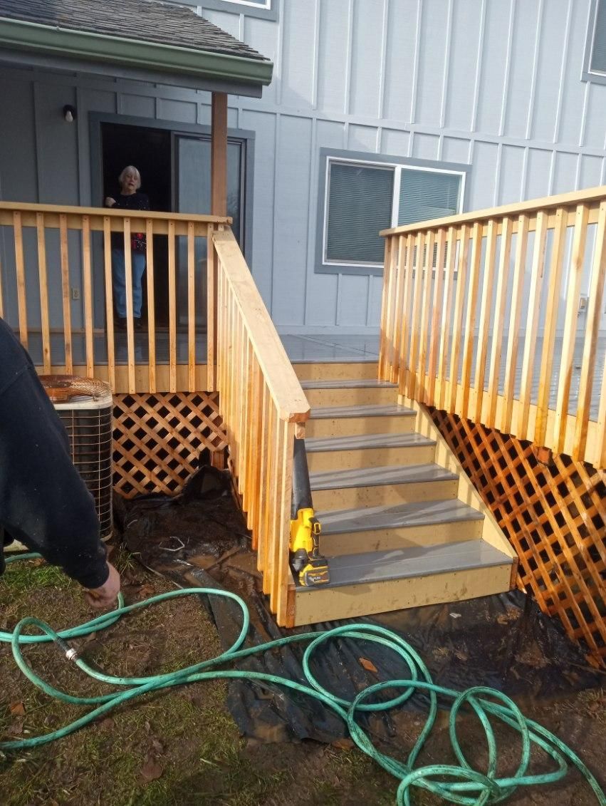 Person hoses a wooden deck with stairs, gray house in background.