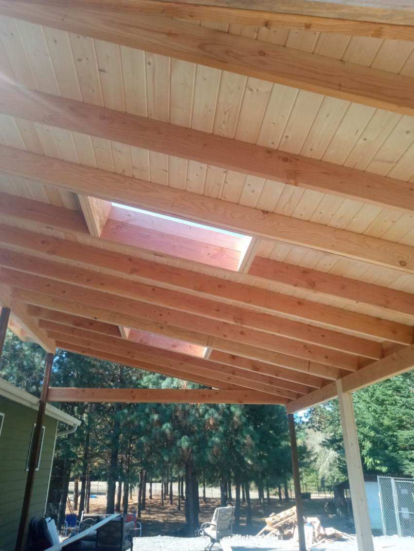 Wooden roof with skylight, supported by posts in an outdoor setting with trees in the background.