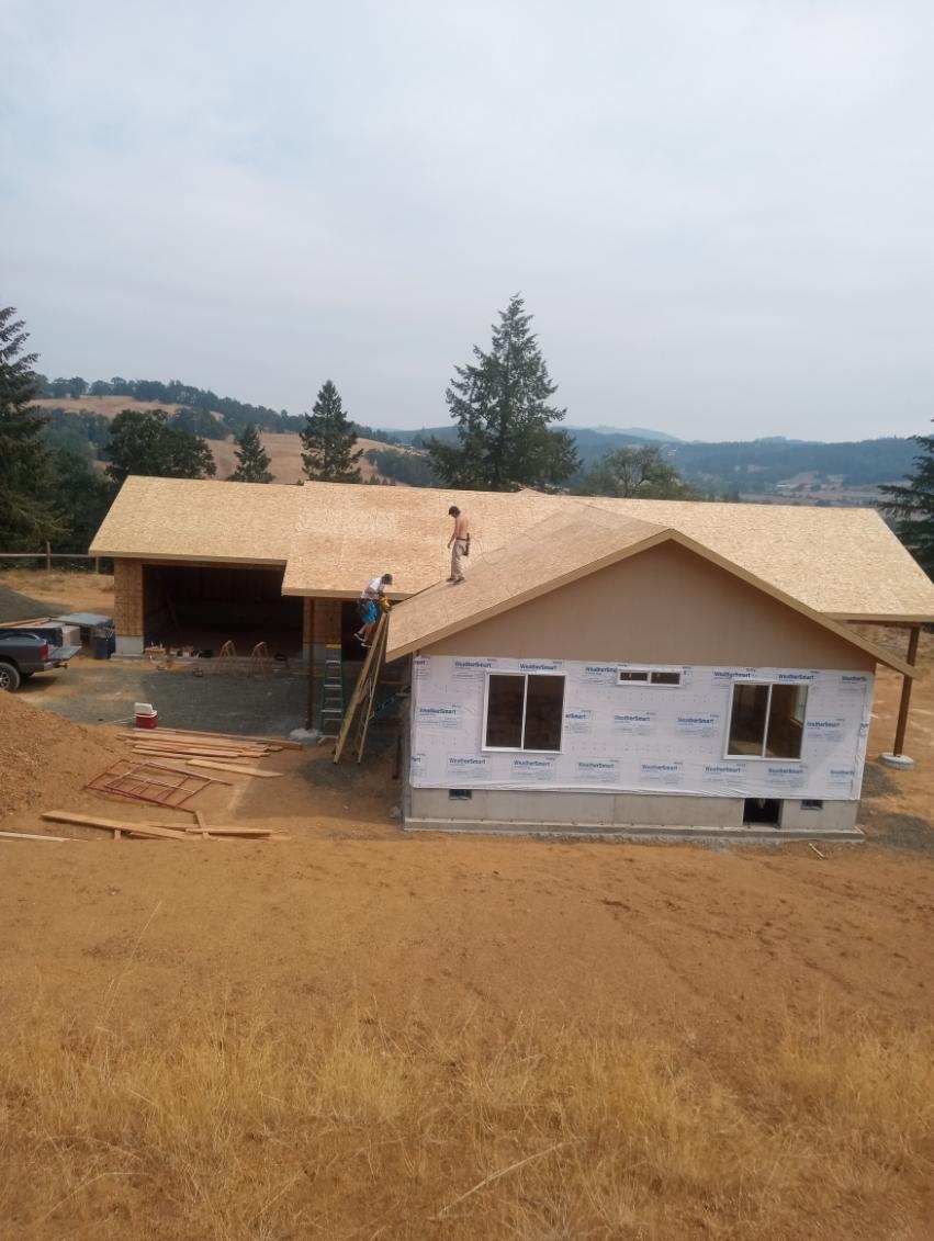House under construction with workers on the roof, in a field, with hills in the background.