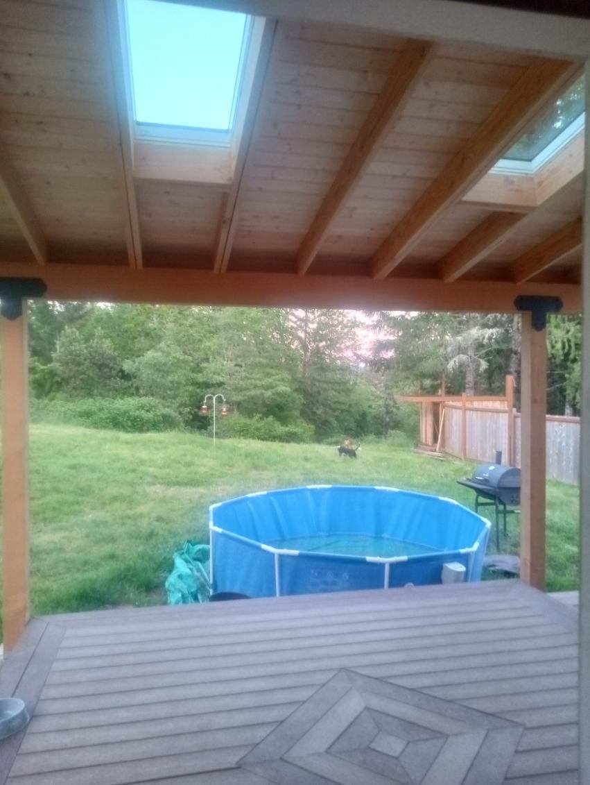 A wooden deck with a blue pool in the backyard, viewed from underneath a wooden roof with skylights.