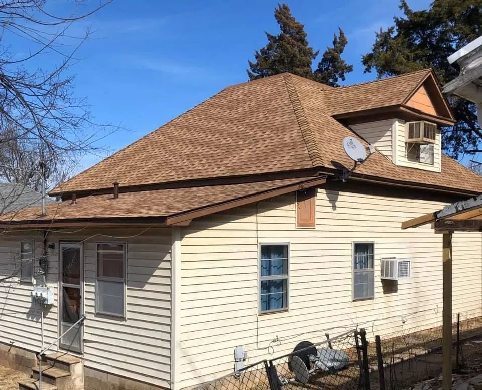 Tan house with brown roof against a clear blue sky. A dormer and satellite dish are visible.